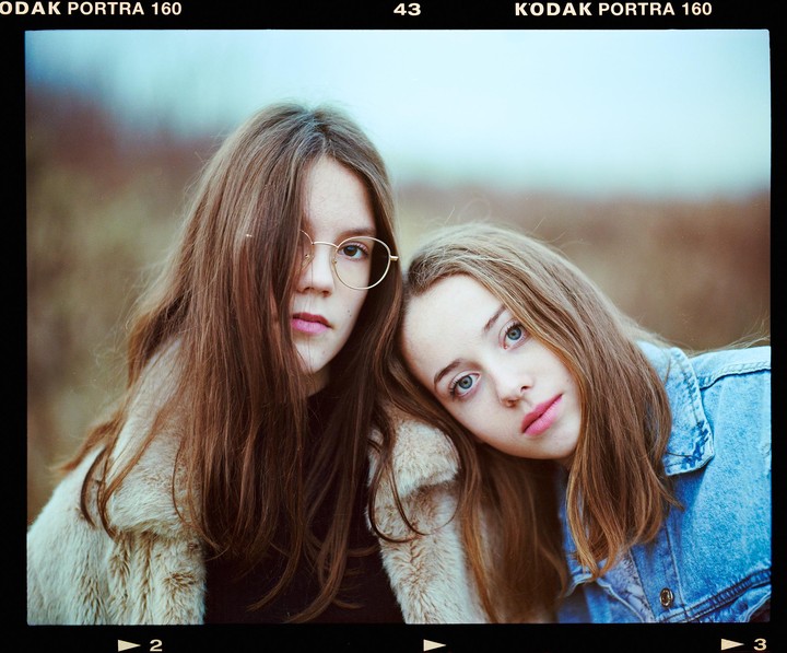 Two girls pose for a portrait, one wearing glasses and a fur coat, the other a denim jacket.