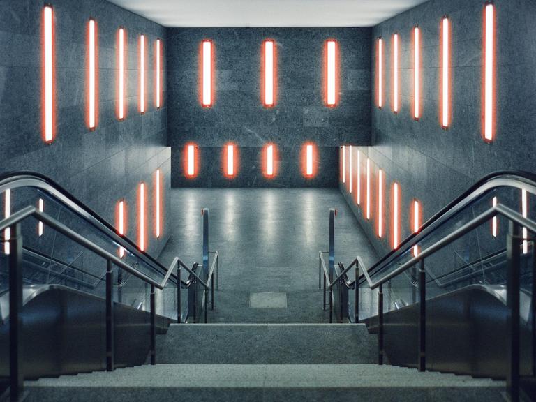 An empty subway station features two escalators descending into a brightly lit underground area with red neon lights on the walls.
