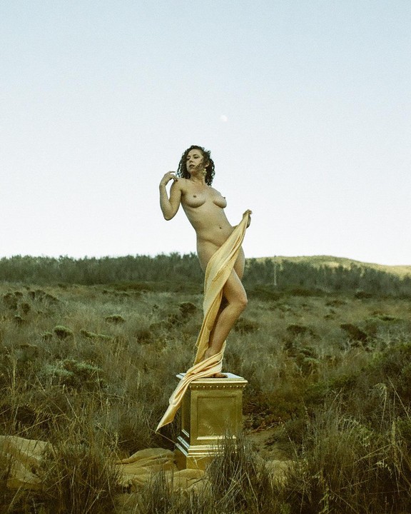A nude woman stands on a golden pedestal in a grassy field, draped with a golden fabric.