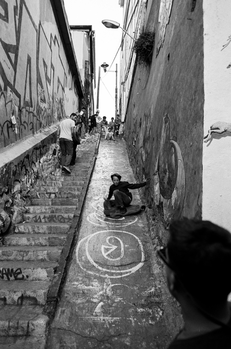 A boy slides down a steep, narrow street with graffiti-covered walls and steps on one side, while other people gather further up the street.