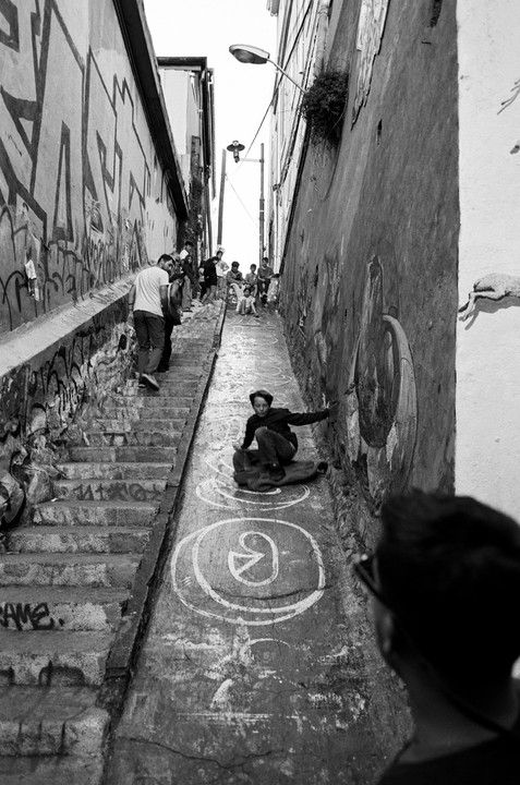 A boy slides down a steep, narrow street with graffiti-covered walls and steps on one side, while other people gather further up the street.