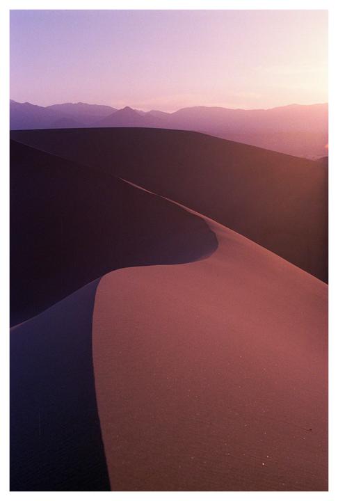 A desert landscape shows sand dunes and mountains under a purple sky at dusk.