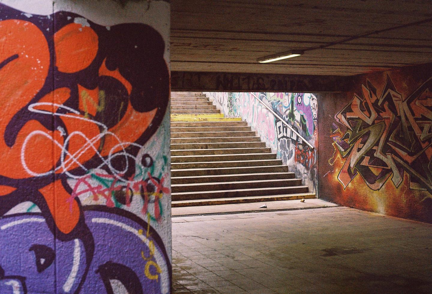 A graffiti-covered underpass leads to a set of stairs with a metal handrail.