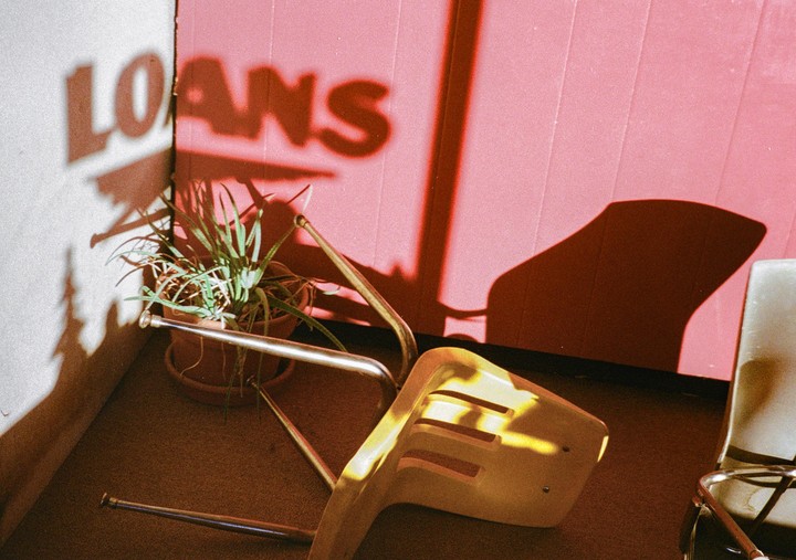 The shadow of the word "LOANS" is cast on a white wall next to a pink wall, a potted plant, and a yellow chair.