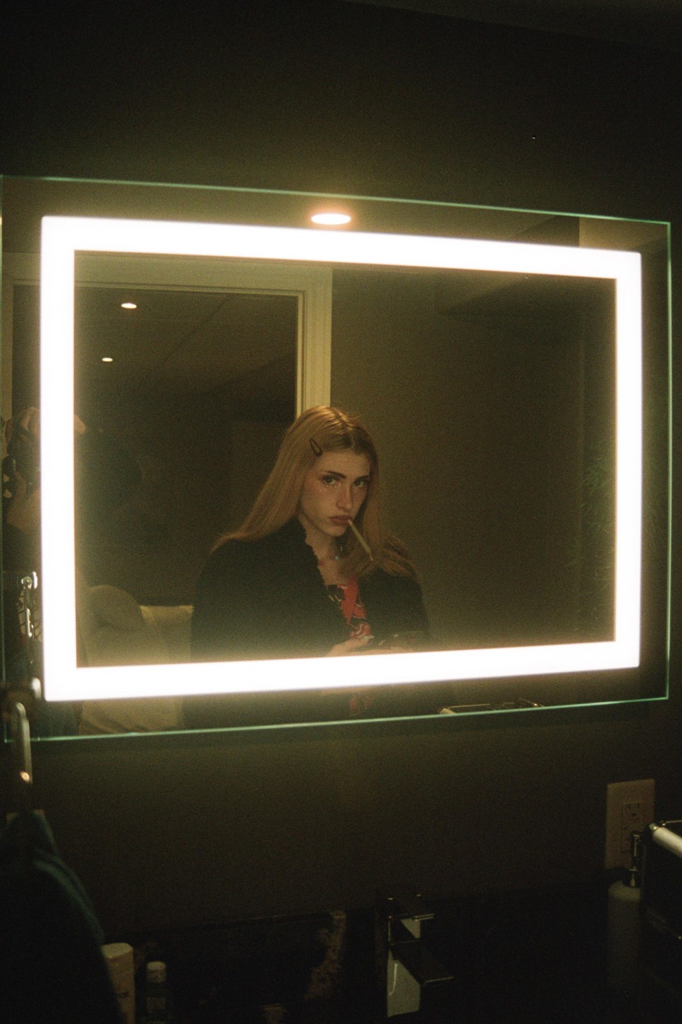 A woman with long hair and hair clips in her hair looks at her reflection in a mirror with a cigarette in her mouth.