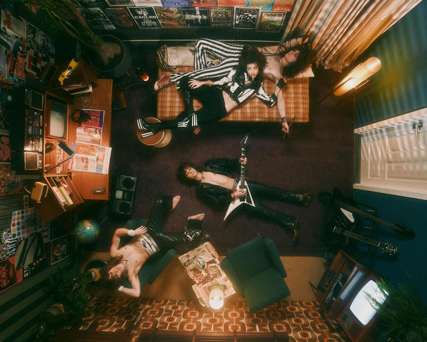 Three musicians are lounging in a cluttered room filled with music posters and instruments.