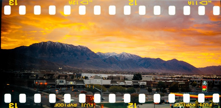 A vibrant orange sunset illuminates a mountain range behind a town with commercial buildings and a street.