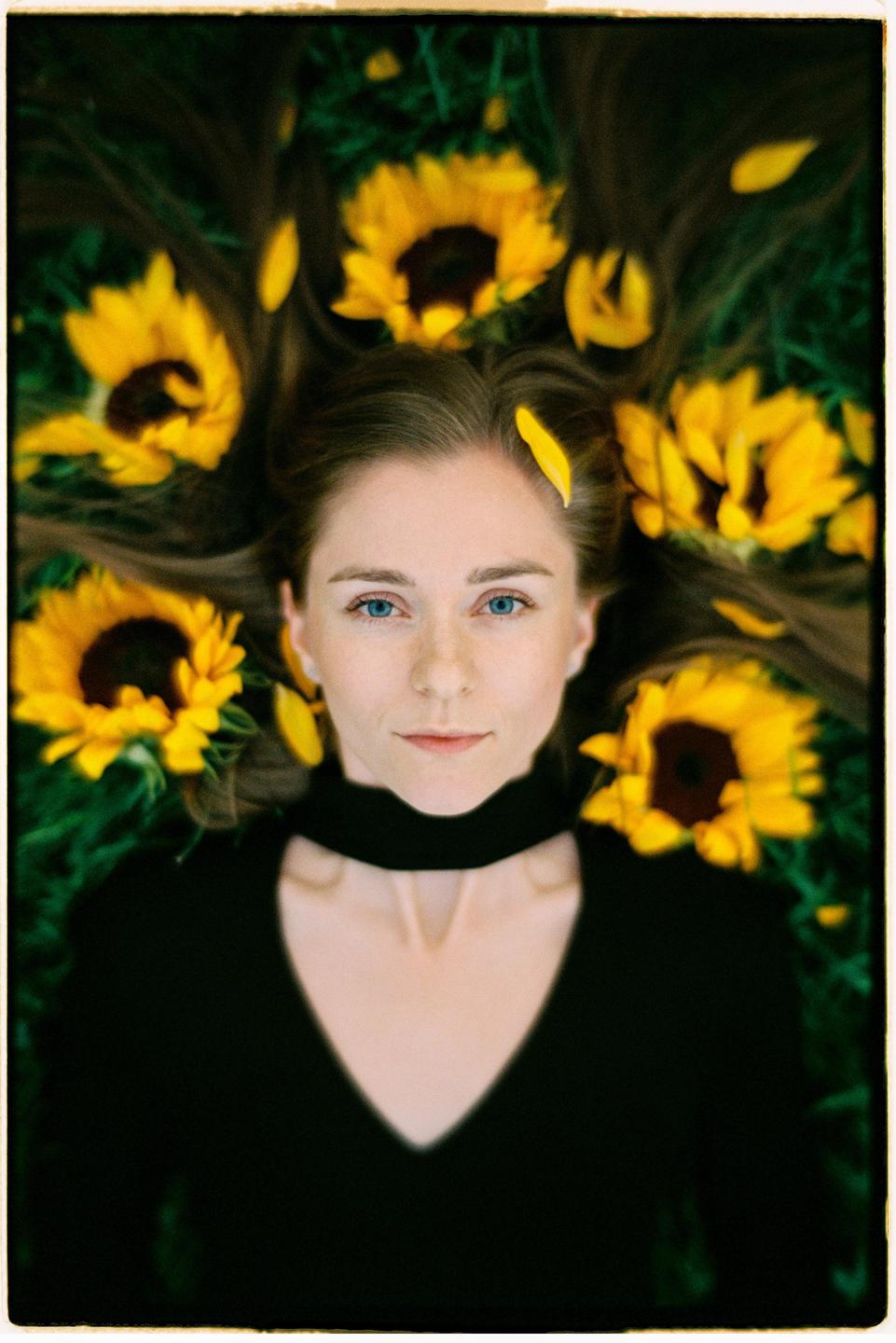 A woman with blue eyes lies on green grass with sunflowers and petals in her hair.