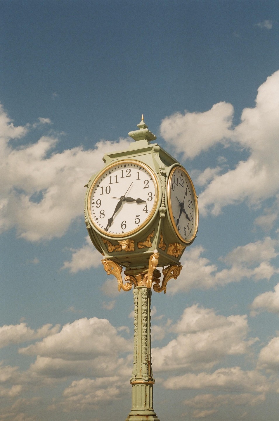 A green and gold ornate street clock stands against a blue sky with white clouds.