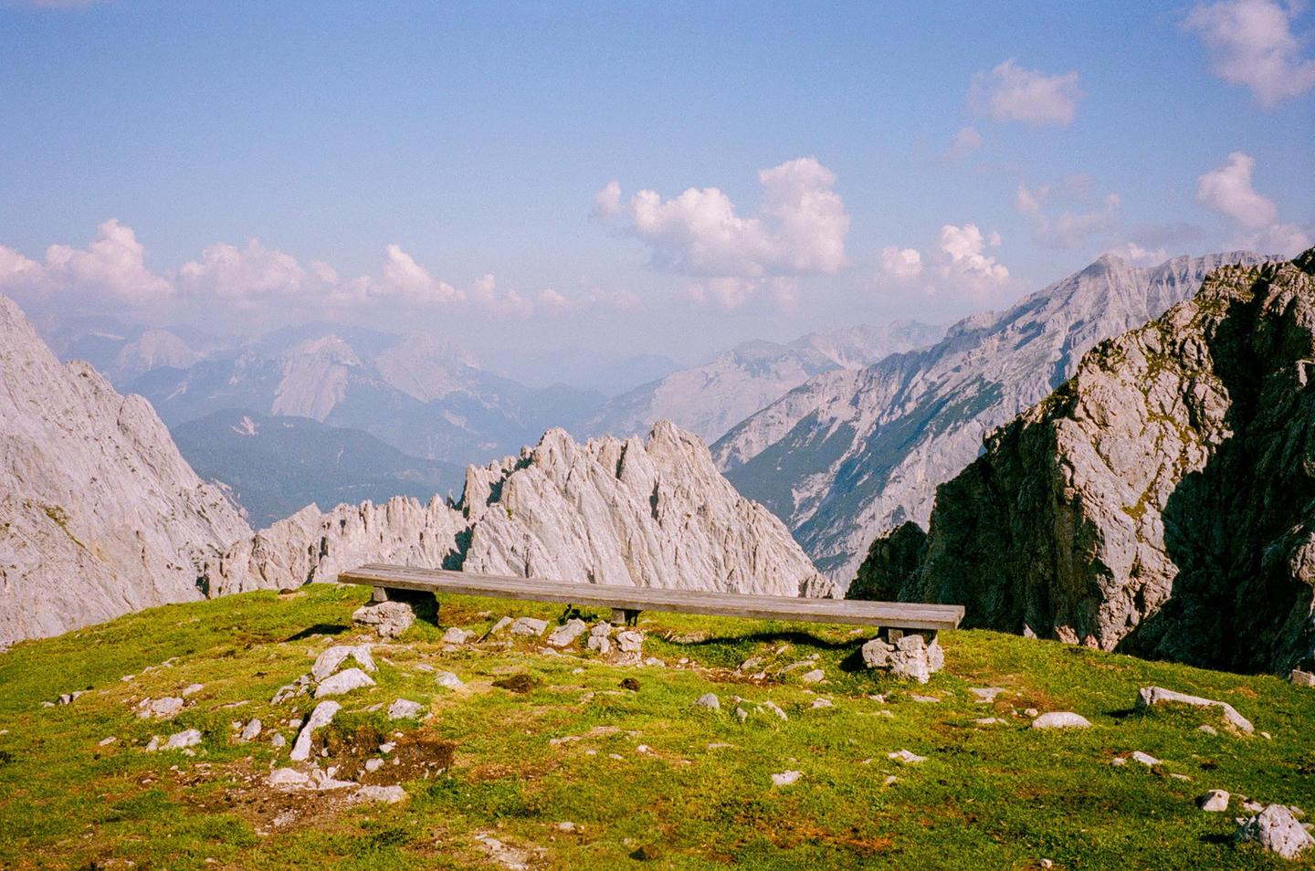A wooden bench sits on a grassy mountain peak overlooking a valley of rocky mountains under a blue sky with clouds.