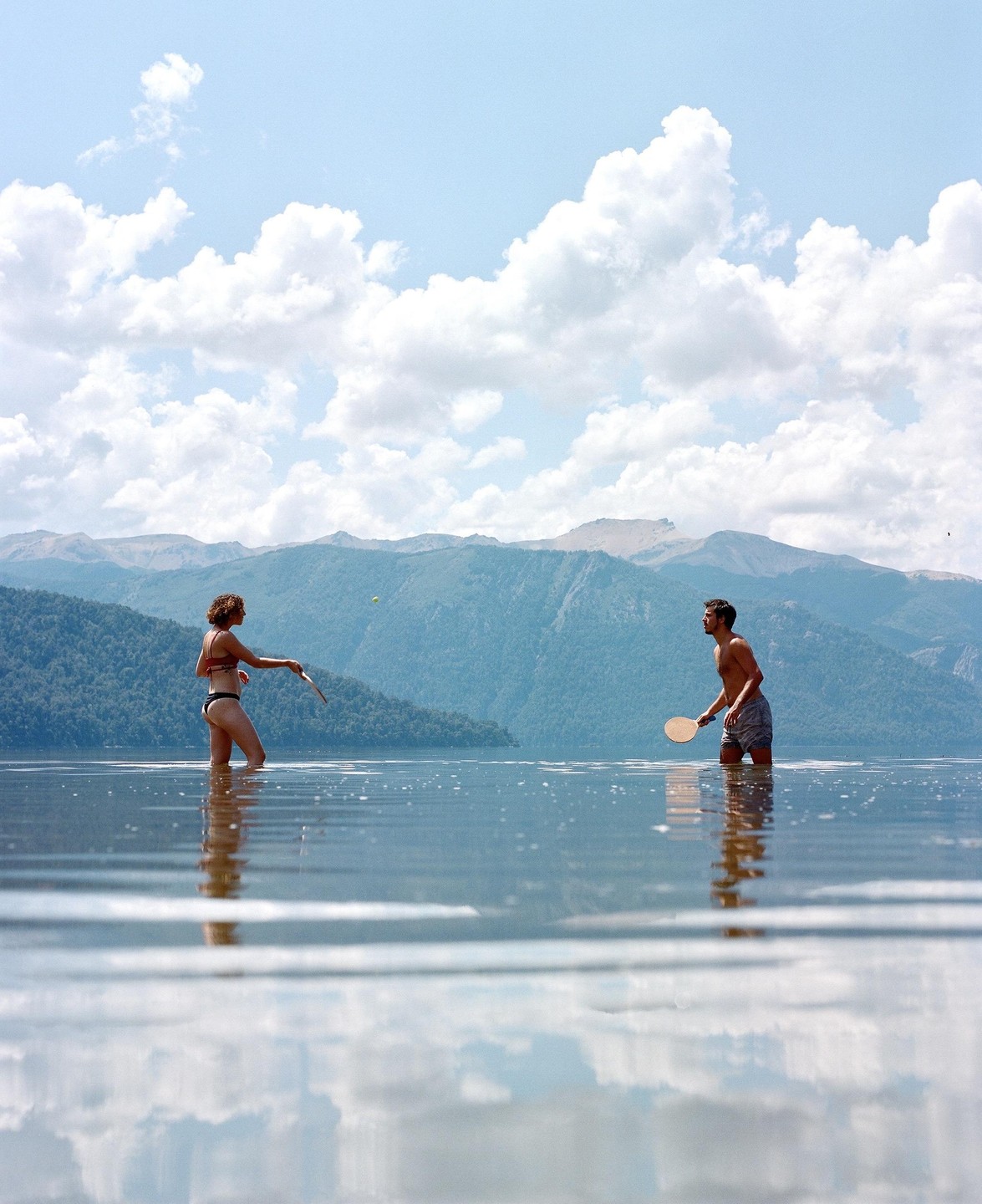 A couple plays paddleball in a lake with mountains in the background.