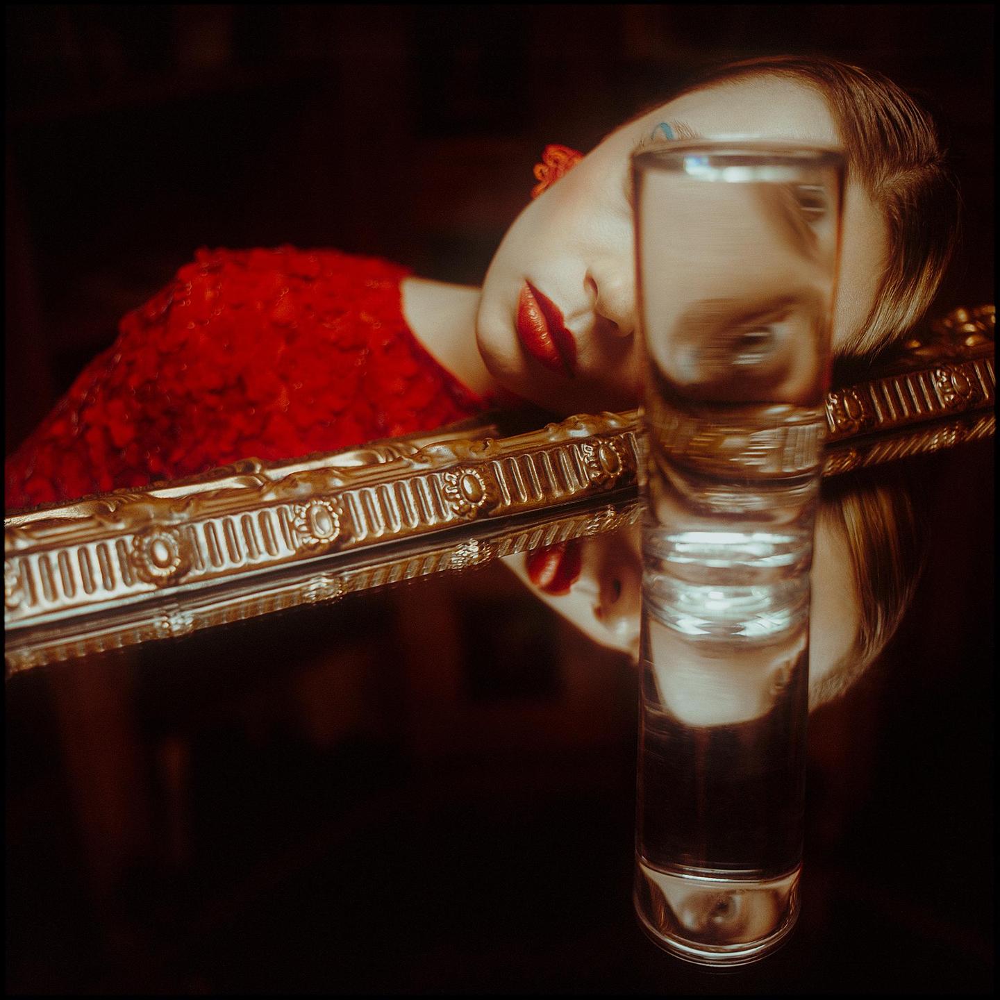 A woman with red lipstick and a red dress is reflected in a mirror, with a glass of water in the foreground distorting her reflection.