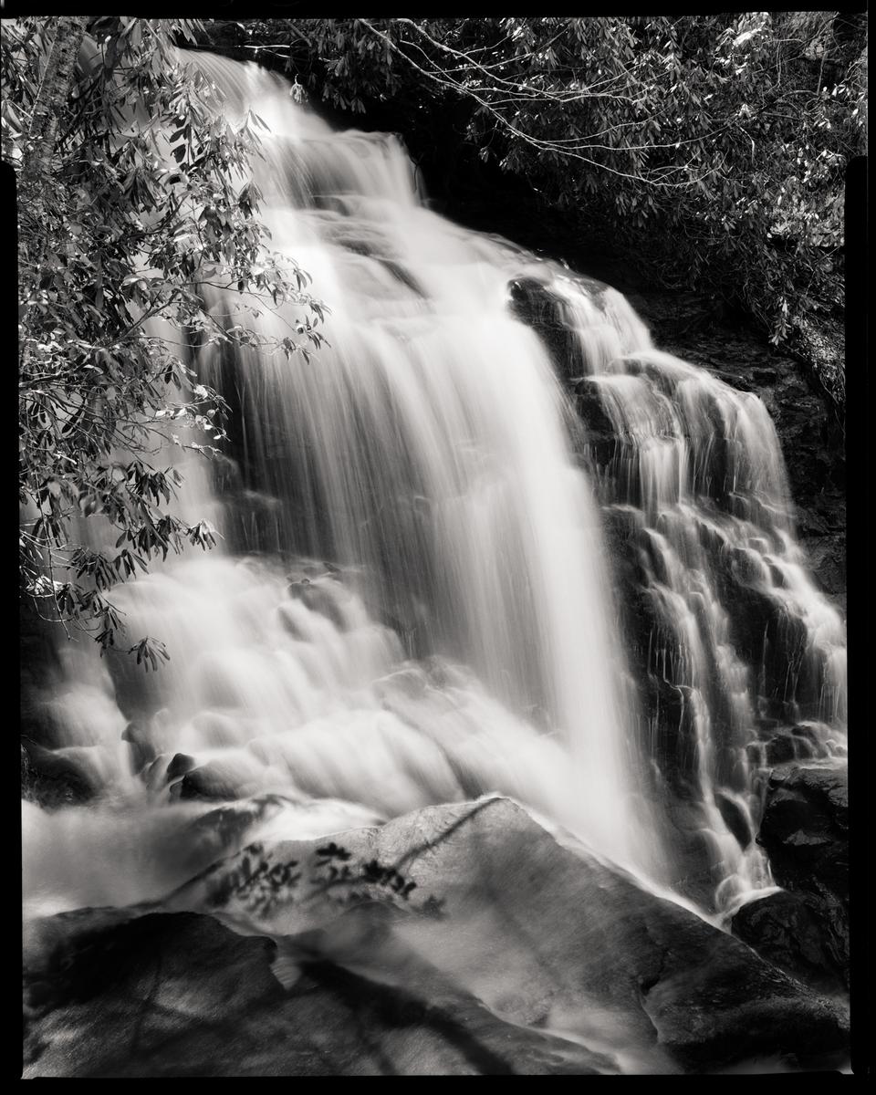 A waterfall cascades over rocks, surrounded by foliage.