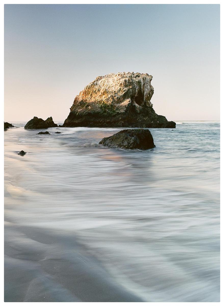 The ocean washes over a sandy beach with several large rocks and a sea stack in the background.