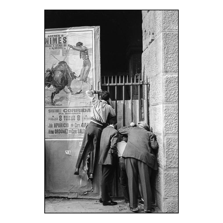 Several men and boys are looking through a barred gate next to a bullfighting poster.