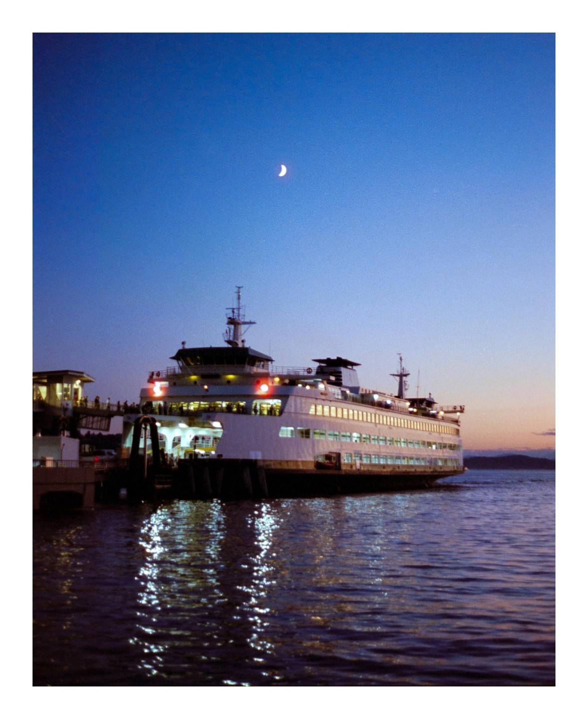 A large ferry boat is docked at a pier under a crescent moon at dusk.