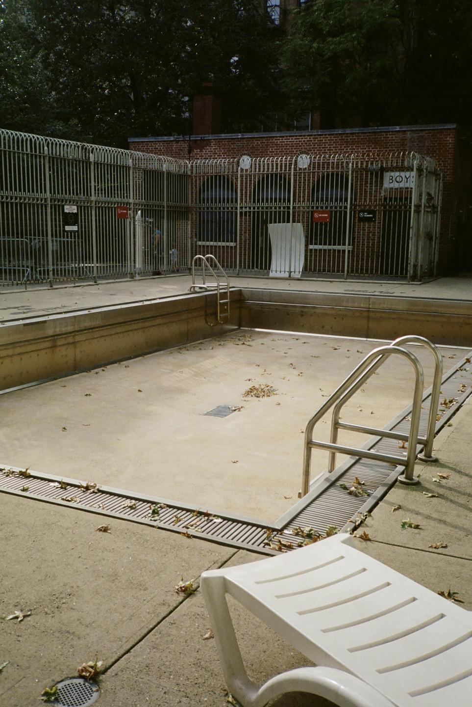 An empty swimming pool with a white lounge chair in the foreground is surrounded by a metal fence and a brick building.