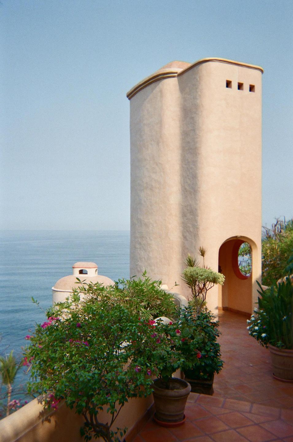 A tall, light-colored tower with an arched doorway stands on a tiled patio overlooking the ocean, surrounded by potted plants.