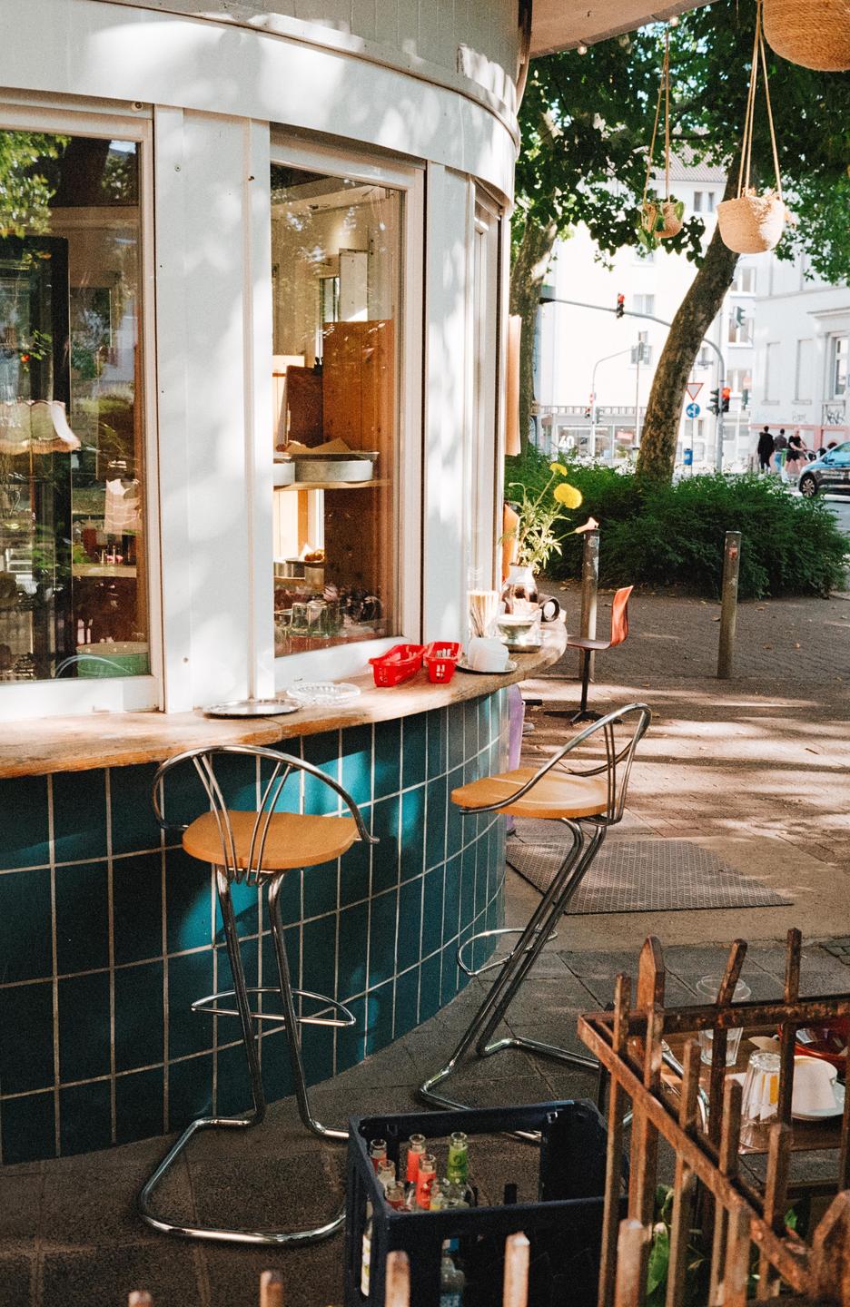 A small outdoor bar with two stools sits on a tiled patio next to a street with trees and buildings.
