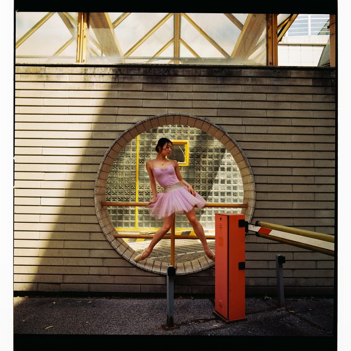 A ballerina in a pink tutu poses in a circular window of a brick building next to a red barrier.
