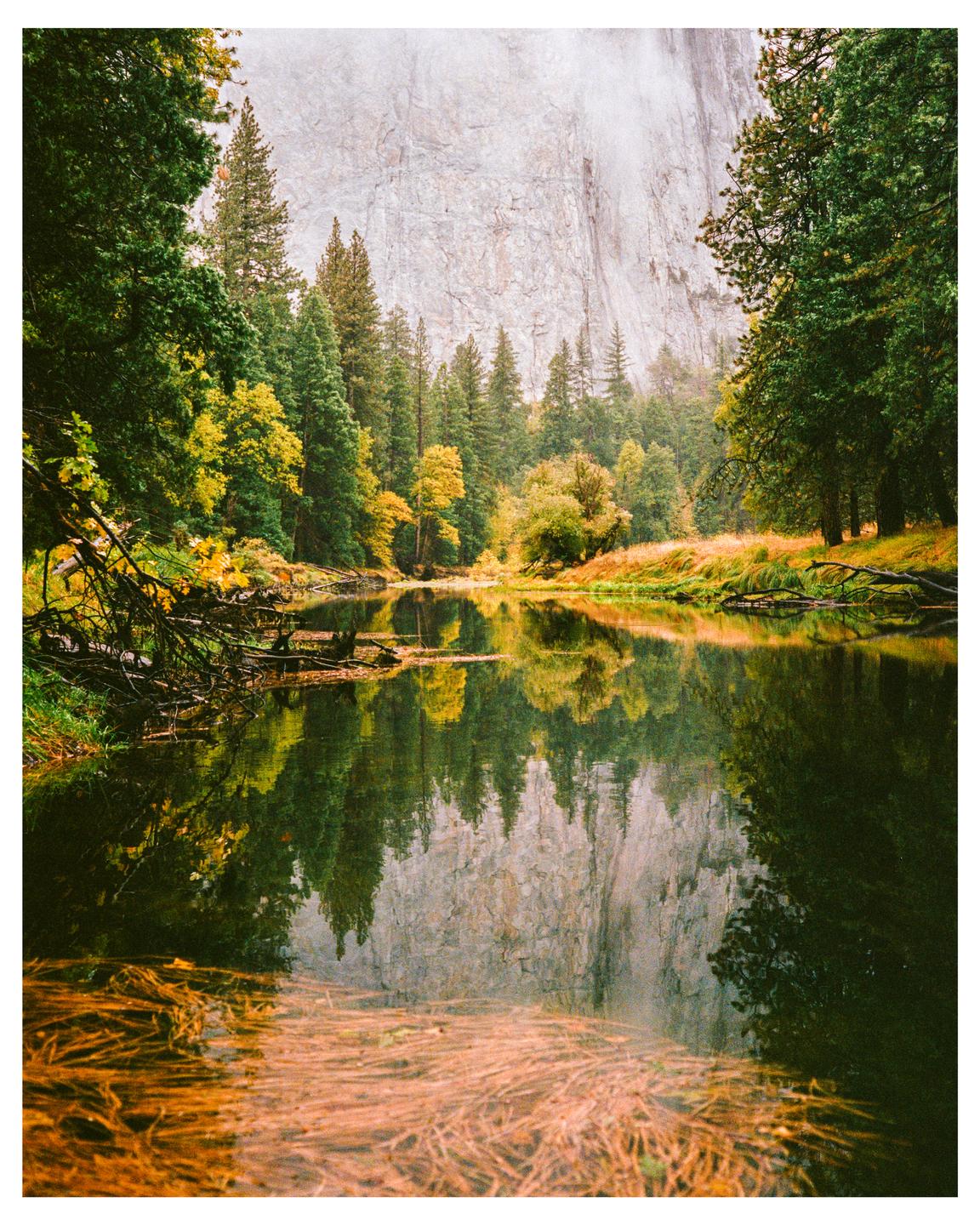 A river reflects a forest and a large rock face under a hazy sky.
