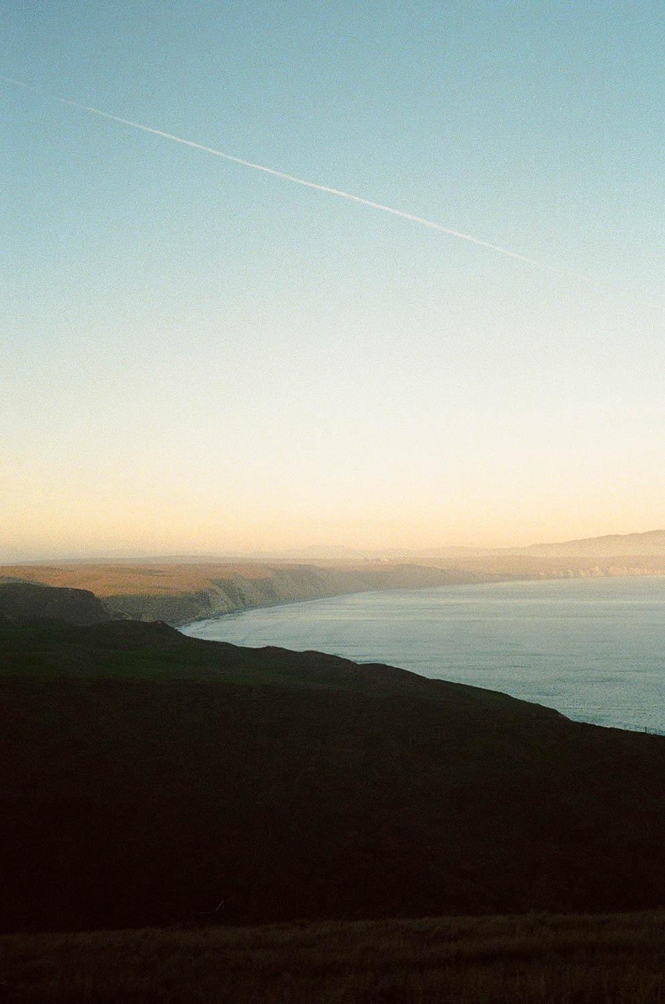 A jet contrail crosses a pale sky above a coastline with dark hills and a calm ocean.
