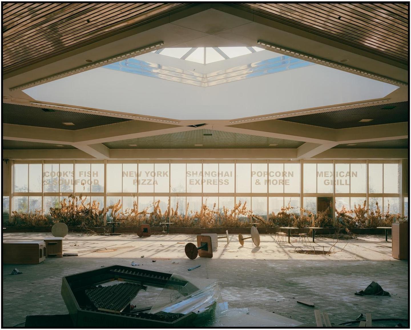An abandoned food court with restaurant names on the windows is illuminated by sunlight from a large skylight.