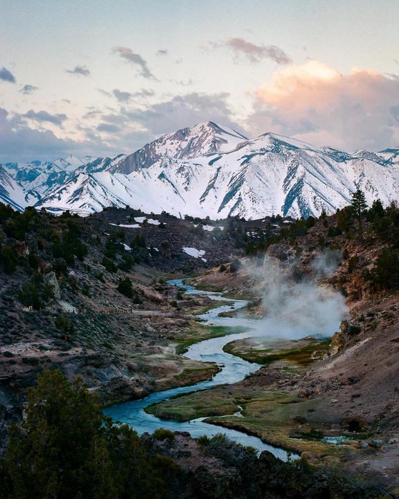 A winding river flows through a valley with steam rising from the bank, leading to snow-capped mountains under a cloudy sky.