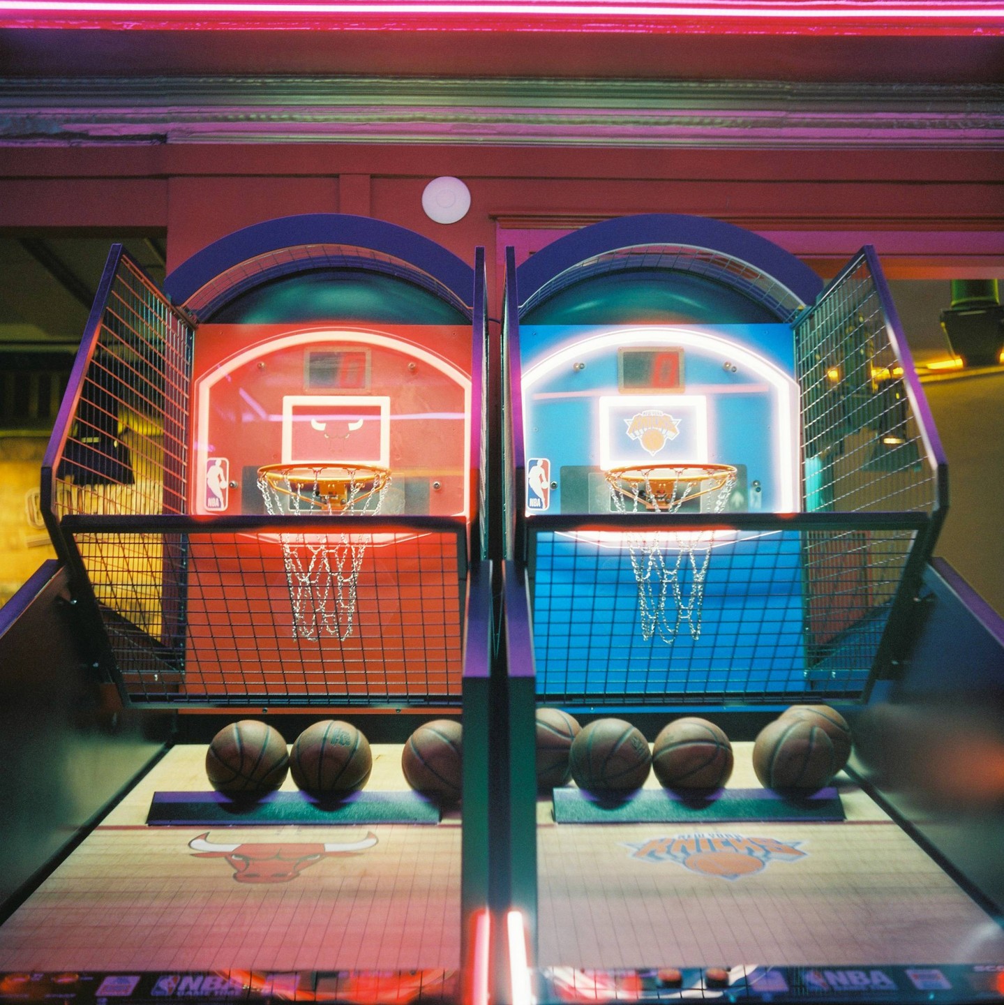 Two basketball arcade games with multiple basketballs are illuminated by neon lights.