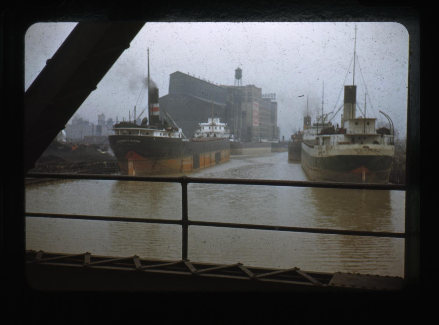 Two freighters are docked in a harbor next to large grain elevators under a hazy sky.