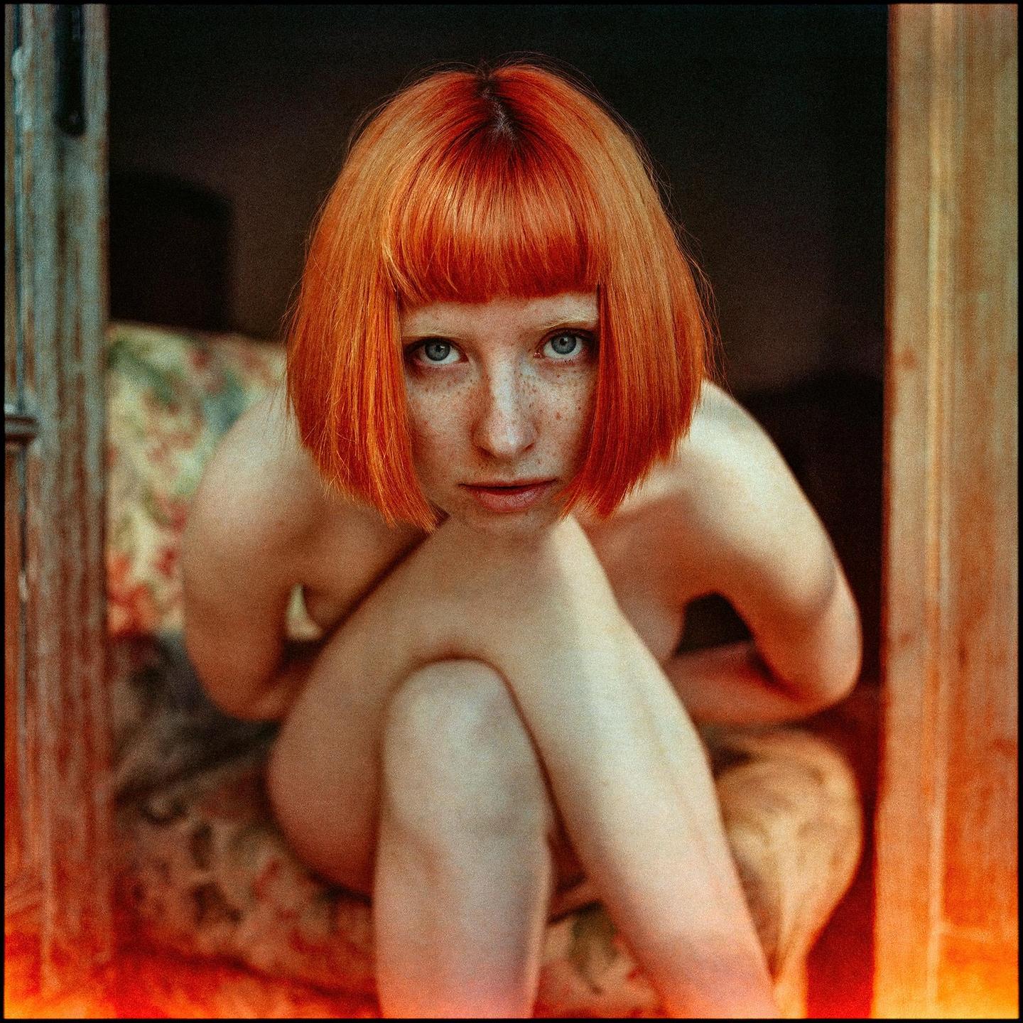 A woman with red hair and freckles sits with her knees up, looking directly at the viewer.