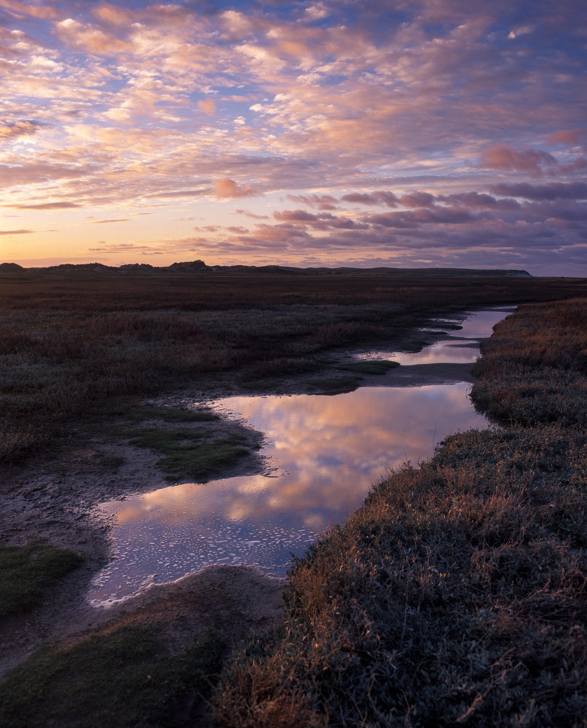 A narrow stream reflects the colorful clouds of a sunset sky in a flat, grassy landscape.