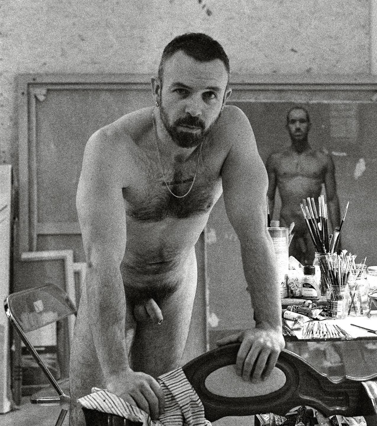 A nude man with a beard leans on a chair in an art studio, with a painting of another nude man in the background.