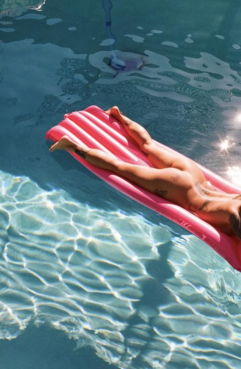 A person with tattoos floats on a pink inflatable raft in a swimming pool.