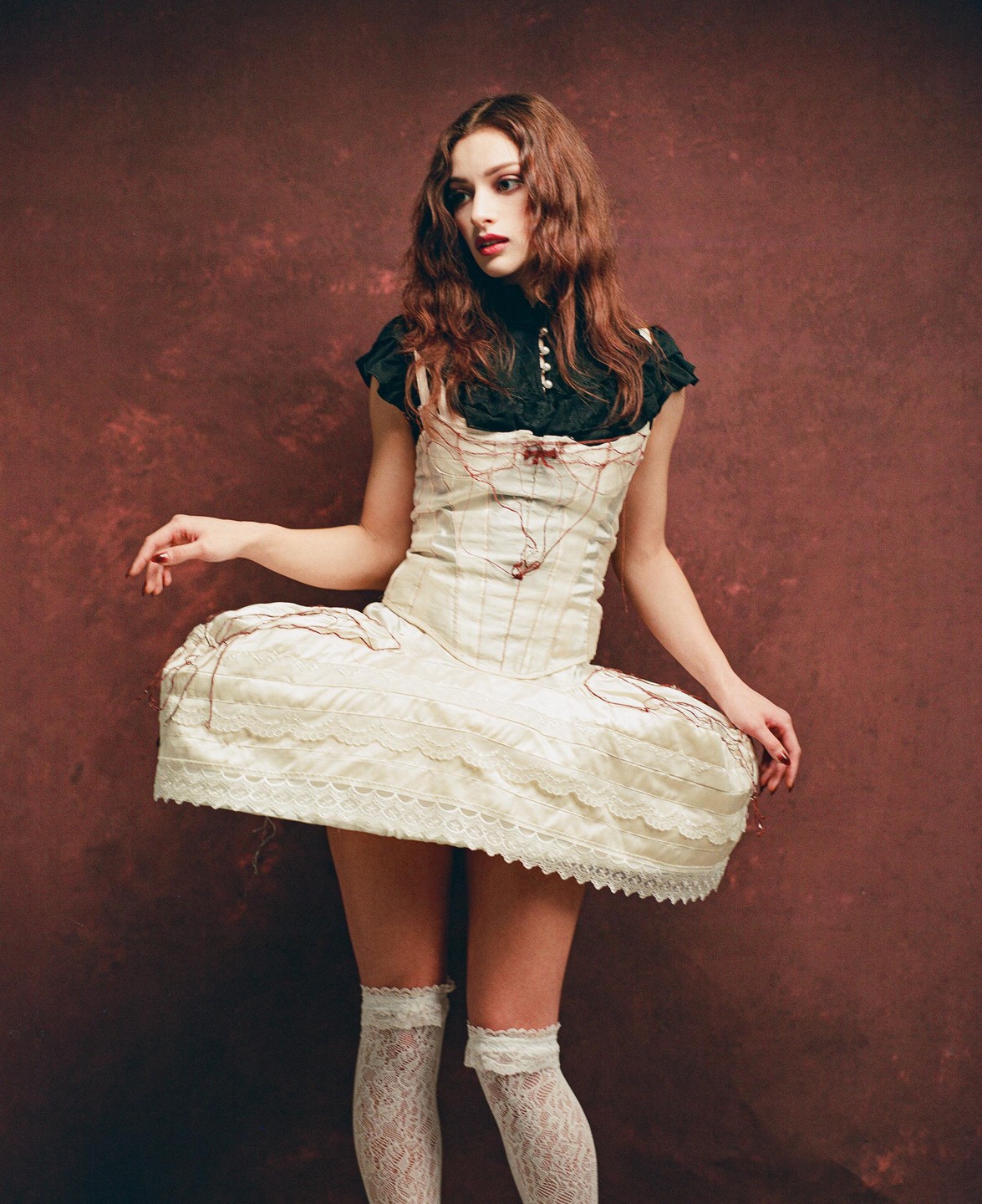 A woman with long, wavy hair poses in a white dress with a black top and white lace stockings against a dark red background.