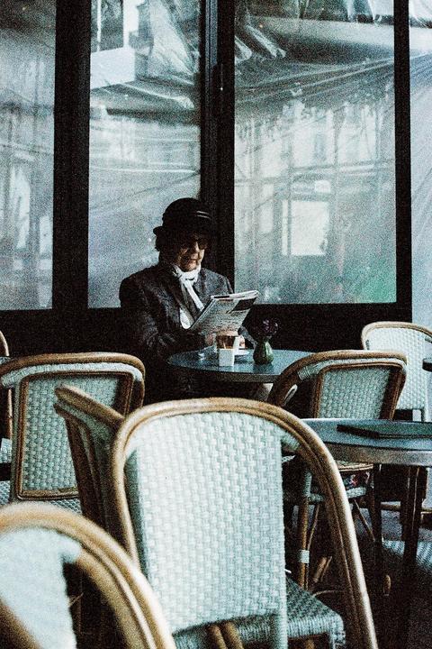 A woman in a hat and scarf reads a newspaper at a cafe table.