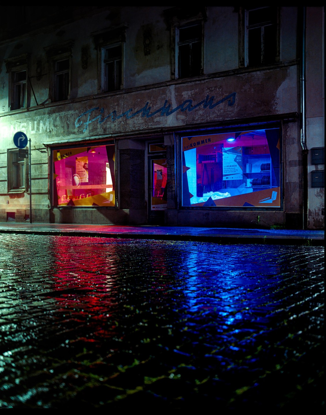A street at night is illuminated by the neon glow of two storefront windows, reflecting on the wet cobblestone.