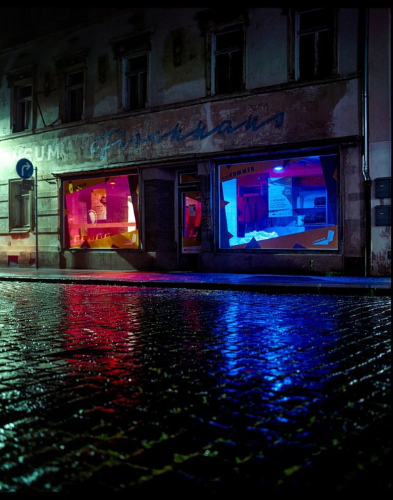A street at night is illuminated by the neon glow of two storefront windows, reflecting on the wet cobblestone.