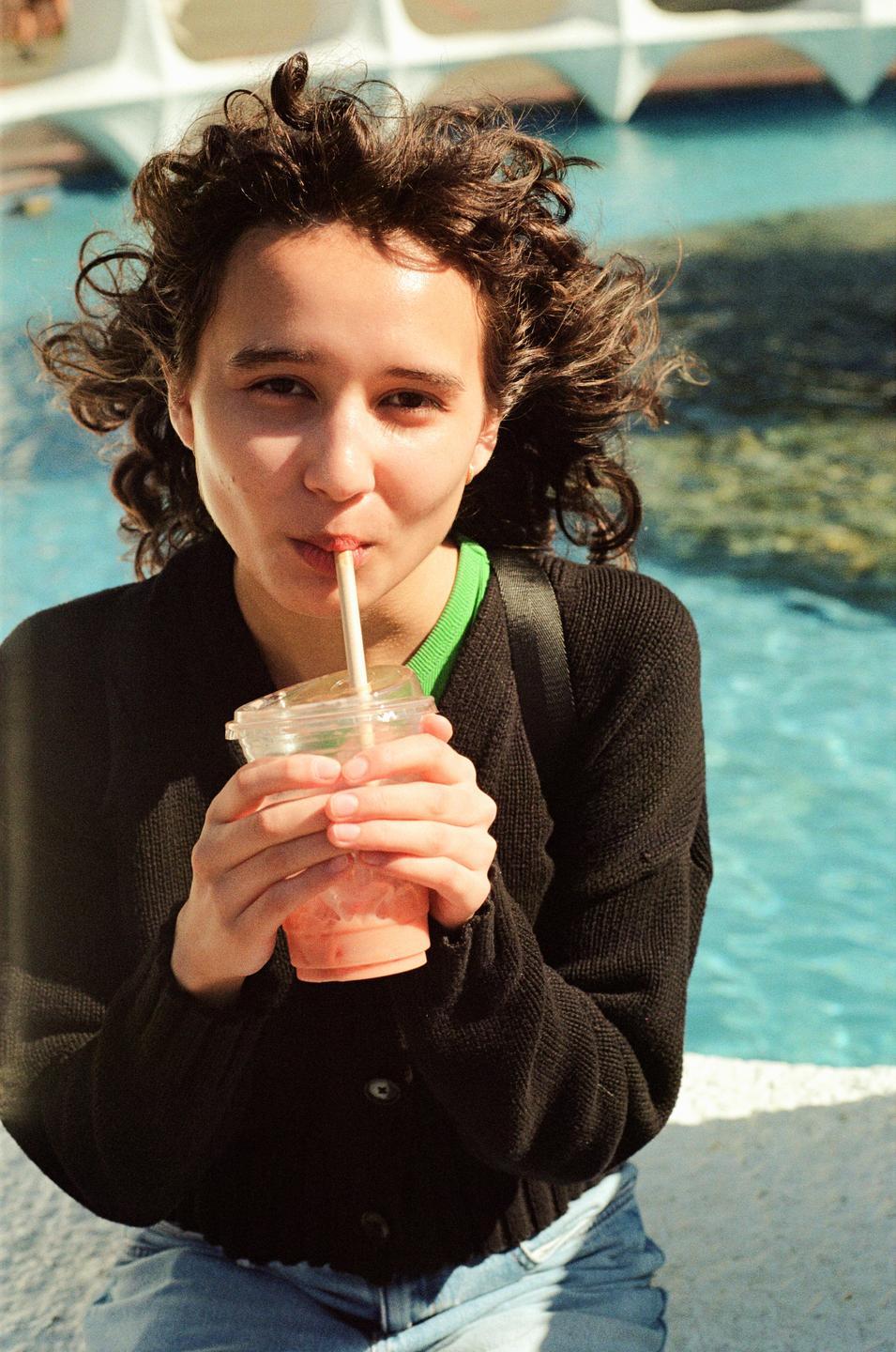 A woman with curly hair sips a drink through a straw, with a body of water and a white structure in the background.