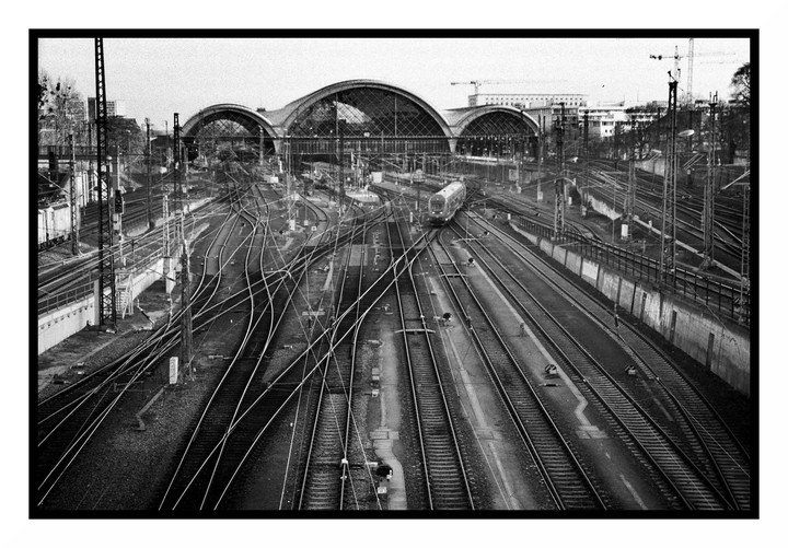 A train station with multiple tracks and an arched glass roof is seen from an elevated perspective.