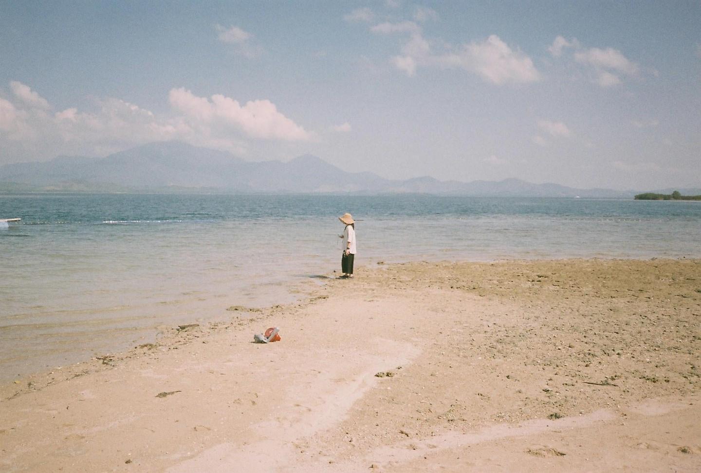 A child wearing a hat stands in shallow water on a sandy beach with mountains in the distance.