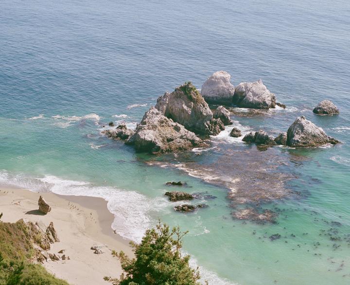 A high-angle view shows a sandy beach with waves breaking, surrounded by rocky outcrops in clear blue and turquoise water.