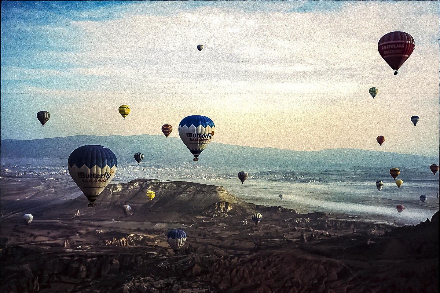 Many hot air balloons float above a vast, rocky landscape with distant mountains under a hazy sky.