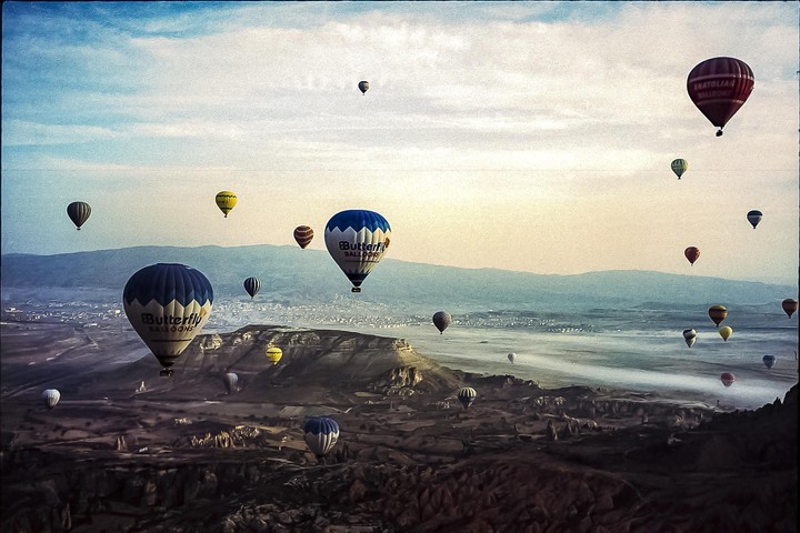 Many hot air balloons float above a vast, rocky landscape with distant mountains under a hazy sky.