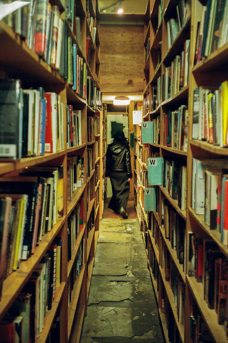 A person in a black leather jacket walks down a narrow aisle between tall bookshelves in a bookstore.