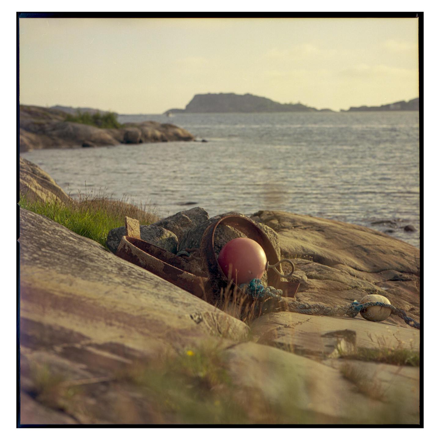 A red buoy and other marine debris rest on rocks by the water with an island in the distance.