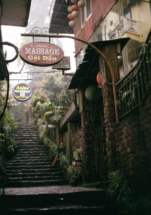 A narrow, steep stone staircase leads up a misty alleyway lined with buildings and lush greenery.