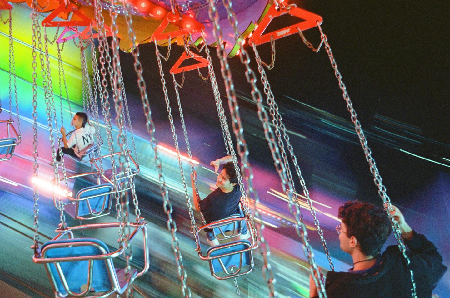 Three people are riding a chain swing ride at night, with colorful light trails in the background.