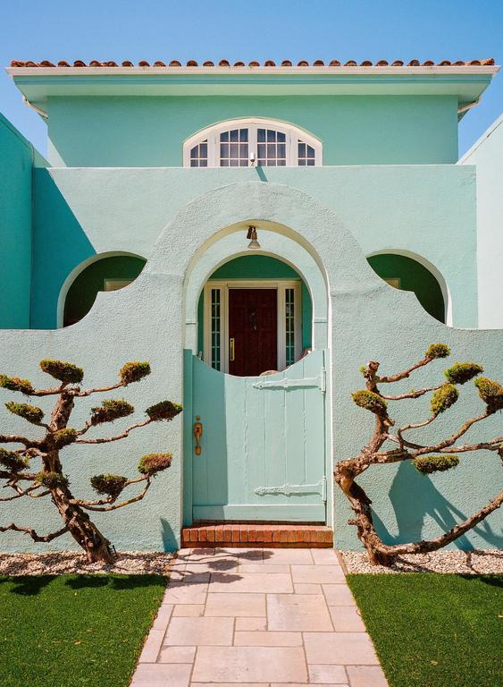 A light blue house with a red tile roof has an arched entryway with a light blue gate and two sculpted trees.