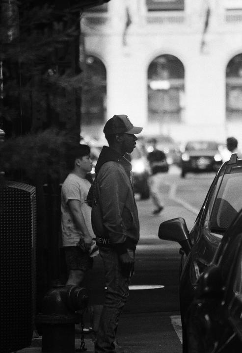 Two men stand on a city street next to a parked car, with a building and other cars in the background.